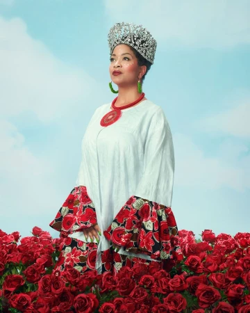 Woman in a white dress and jeweled crown stands among red roses against a blue sky with white clouds, posing in a vibrant setting.